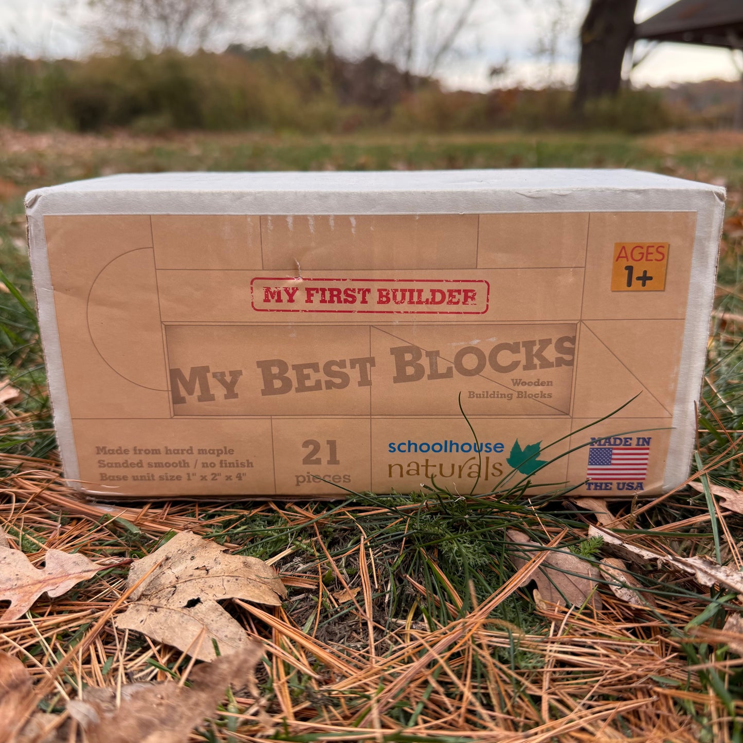 Cardboard box of 'My First Builder My Best Blocks' wooden building blocks by Schoolhouse Naturals on a grass surface.