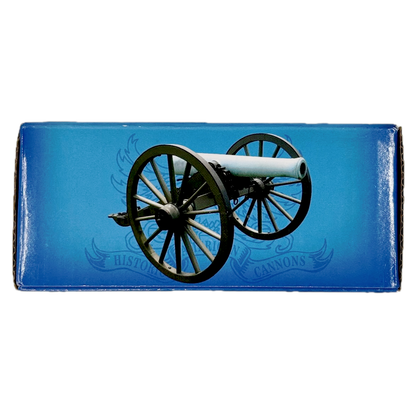 Cardboard box containing small brass replica Trail Cannon toy on a white background