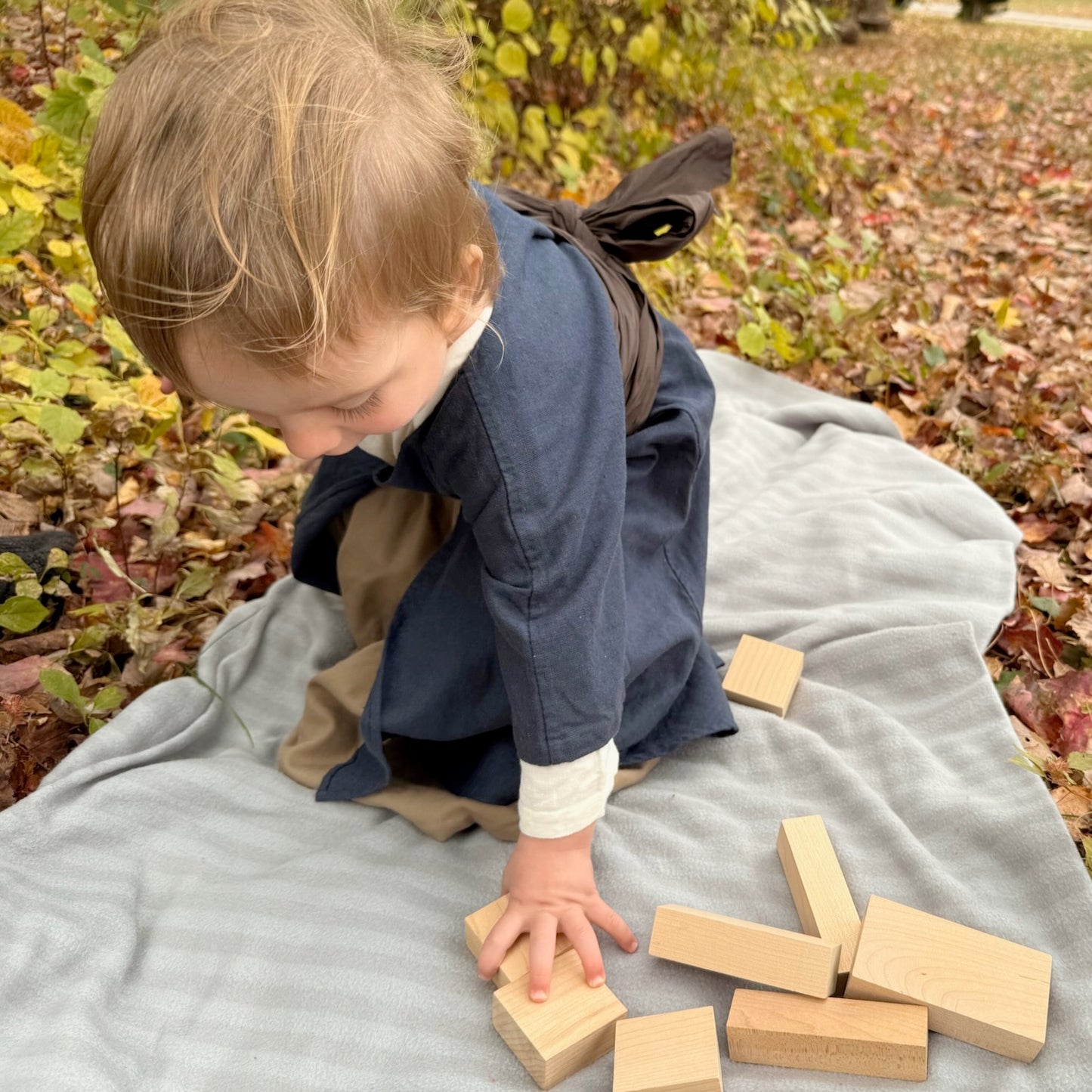 Child playing with wooden blocks on a blanket outdoors in autumn while wearing blue colonial attire.