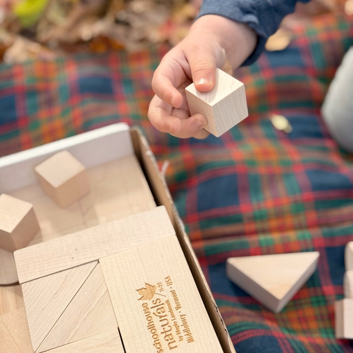 Child's hand holding a wooden block over a box of wooden building blocks with a plaid background.