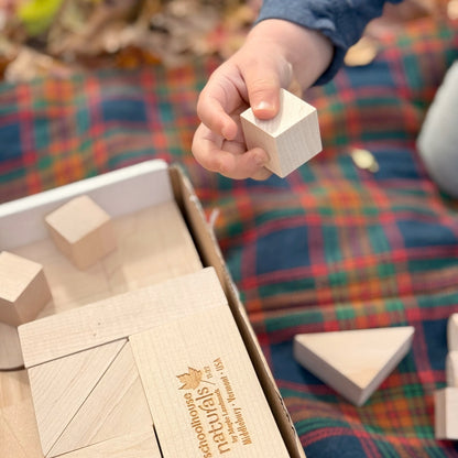 Child's hand holding a wooden block over a box of wooden building blocks with a plaid background.