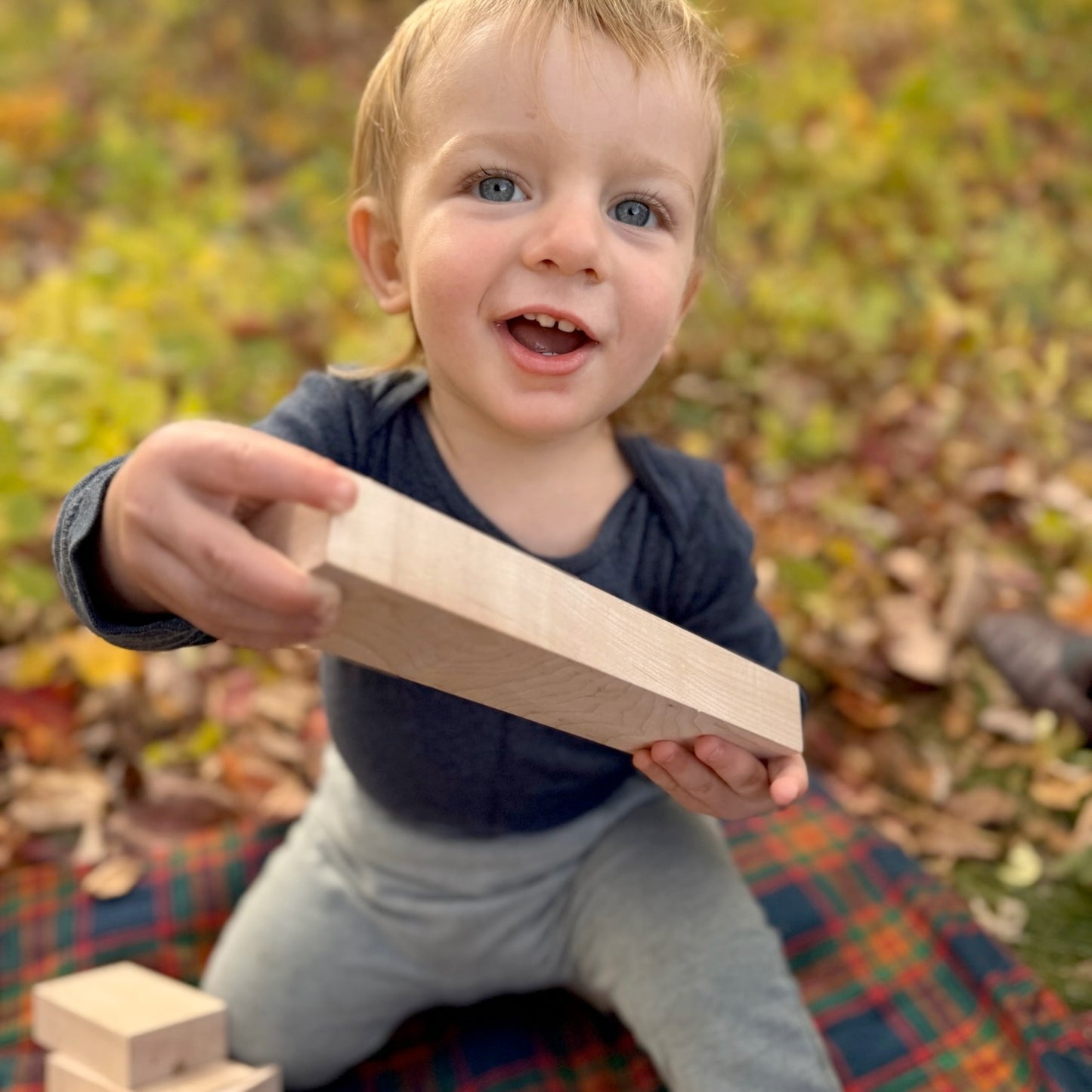Child holding a wooden block outdoors with a colorful blanket and autumn leaves in the background