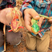 Two children holding colorful toy wooden snakes outdoors on a leafy ground.