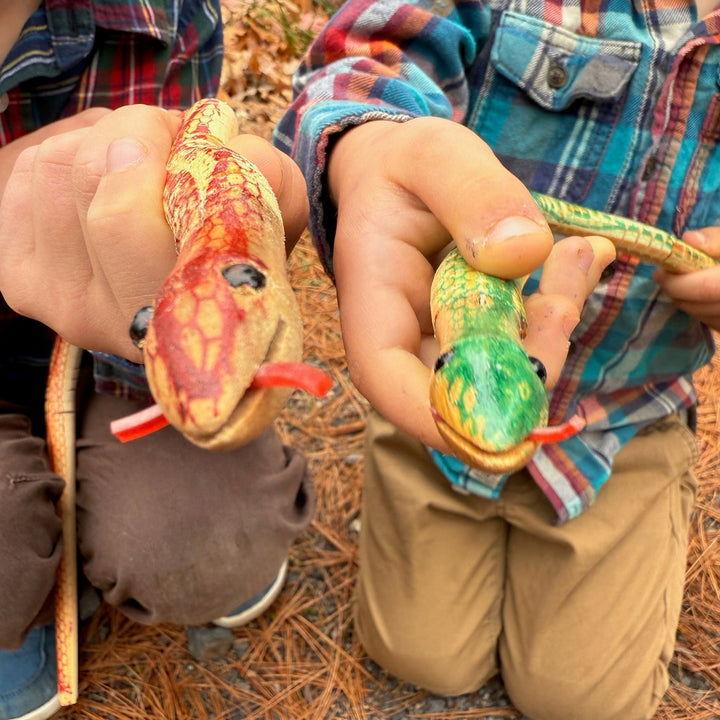 Two children holding colorful toy wooden snakes outdoors on a leafy ground.