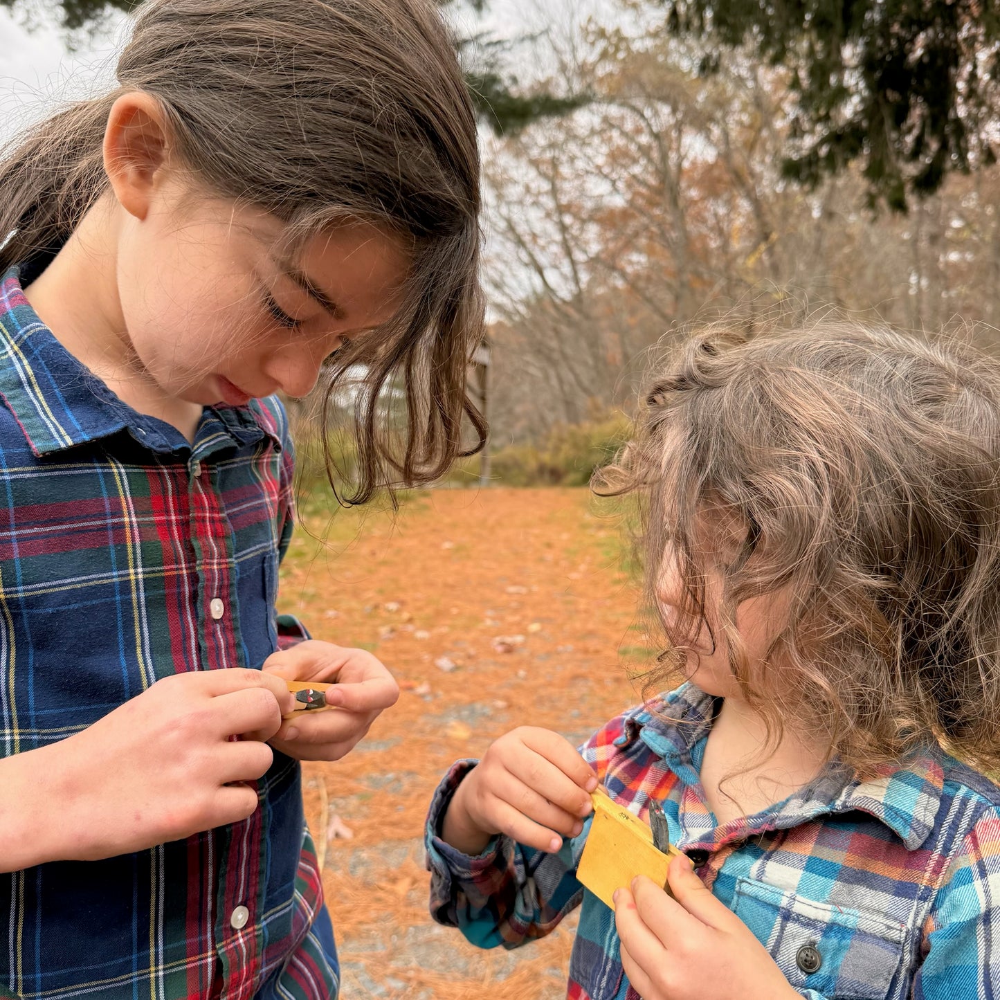 Two children in plaid shirts outdoors, playing with small wooden Snake in the Box toys.