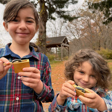 Two children in plaid shirts holding small wooden Snake in the Box toys outdoors with trees in the background