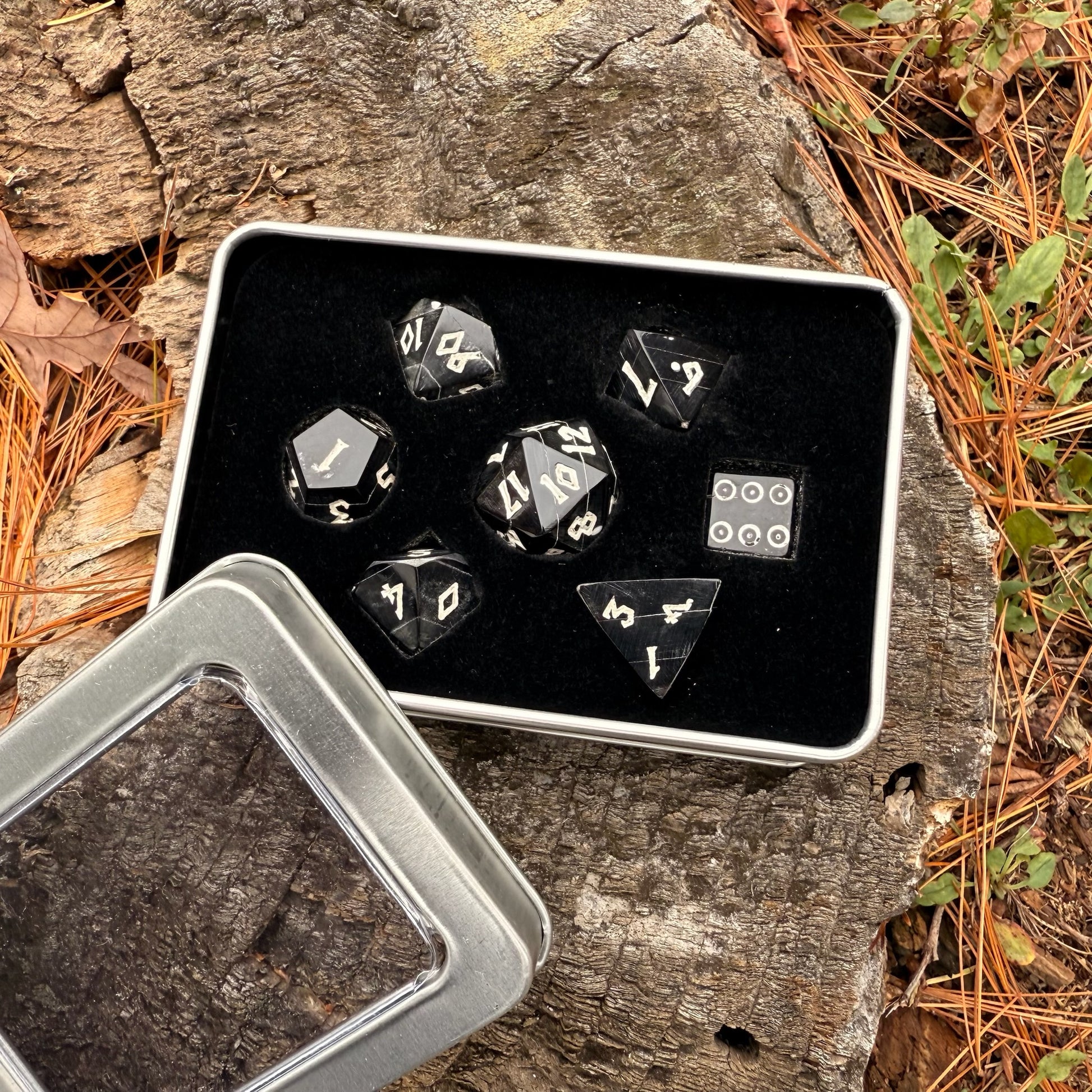 Set of black polyhedral horn dice with white numbers in a metal tin on a wooden surface with leaves.
