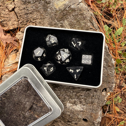 Set of black polyhedral horn dice with white numbers in a metal tin on a wooden surface with leaves.