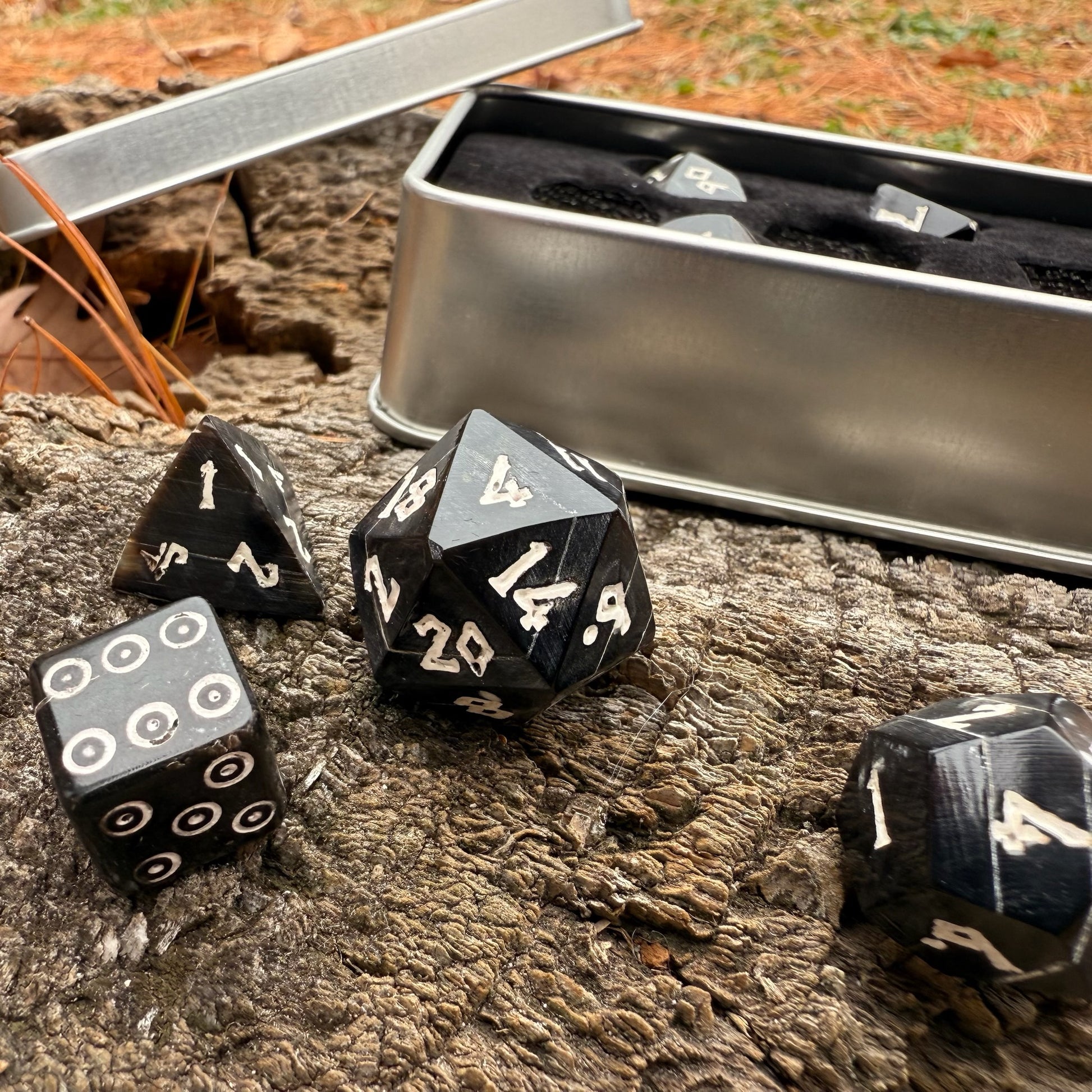 Black polyhedral horn dice with white numbers on a wooden surface next to a metal dice box.