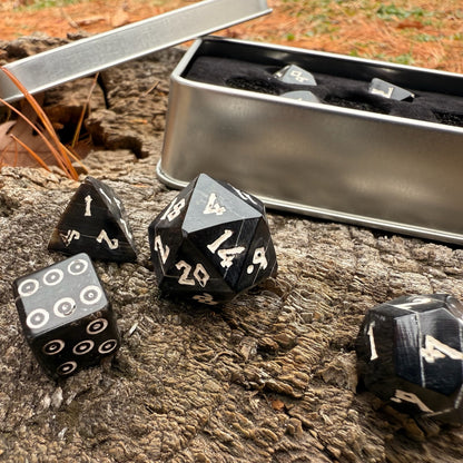 Black polyhedral horn dice with white numbers on a wooden surface next to a metal dice box.