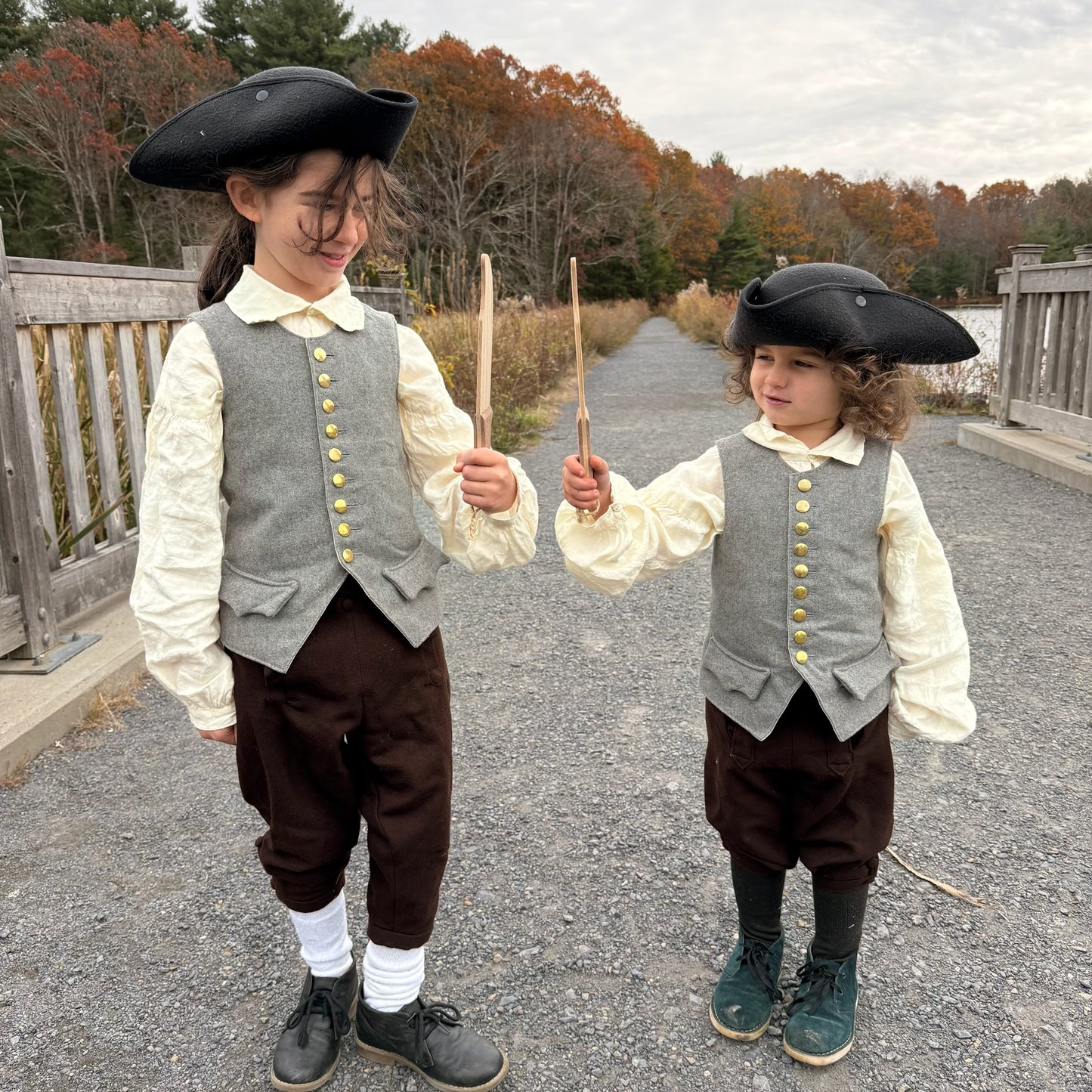 Two children in colonial-style clothing holding wooden toy knives outdoors.