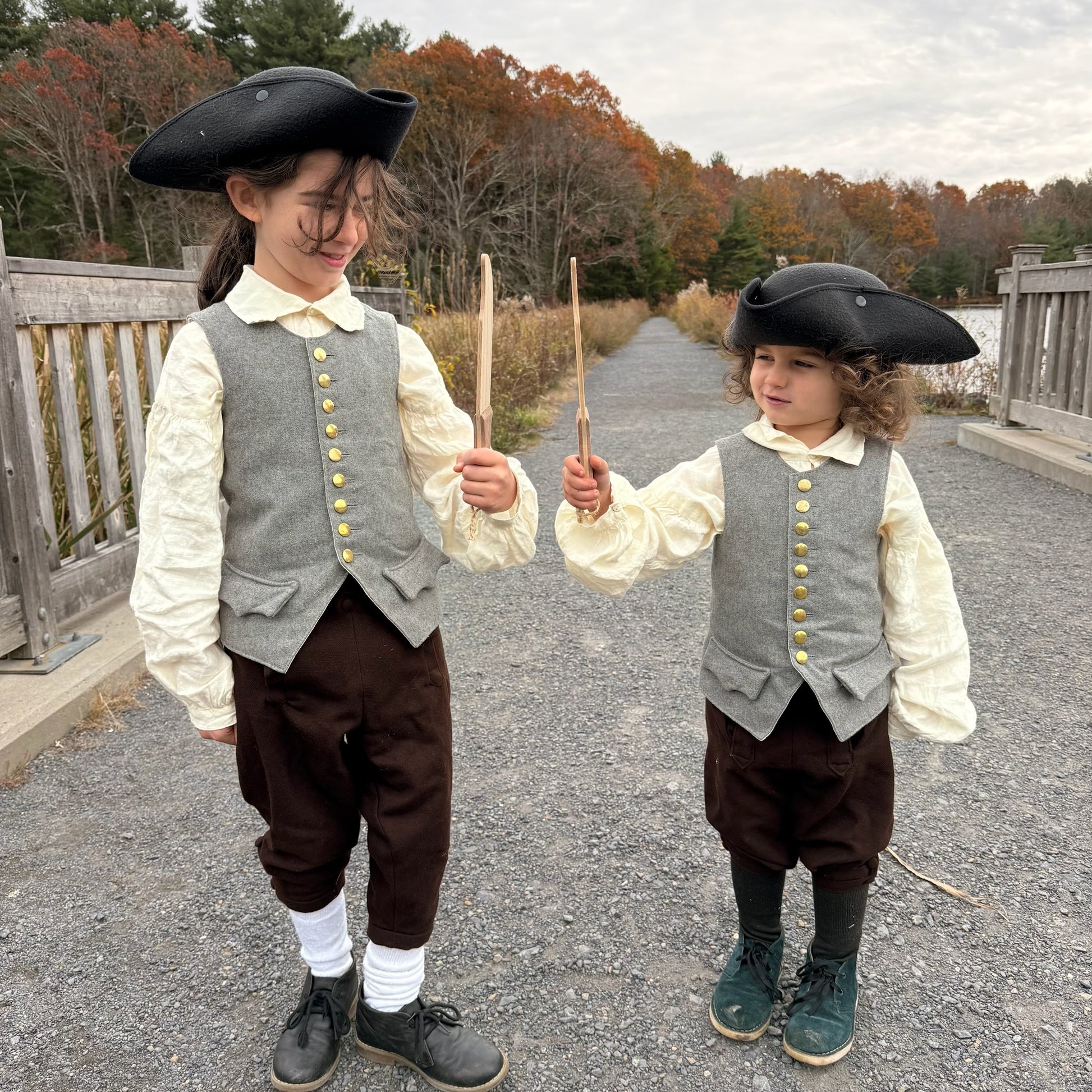 Two children in colonial-style clothing holding wooden toy knives outdoors.