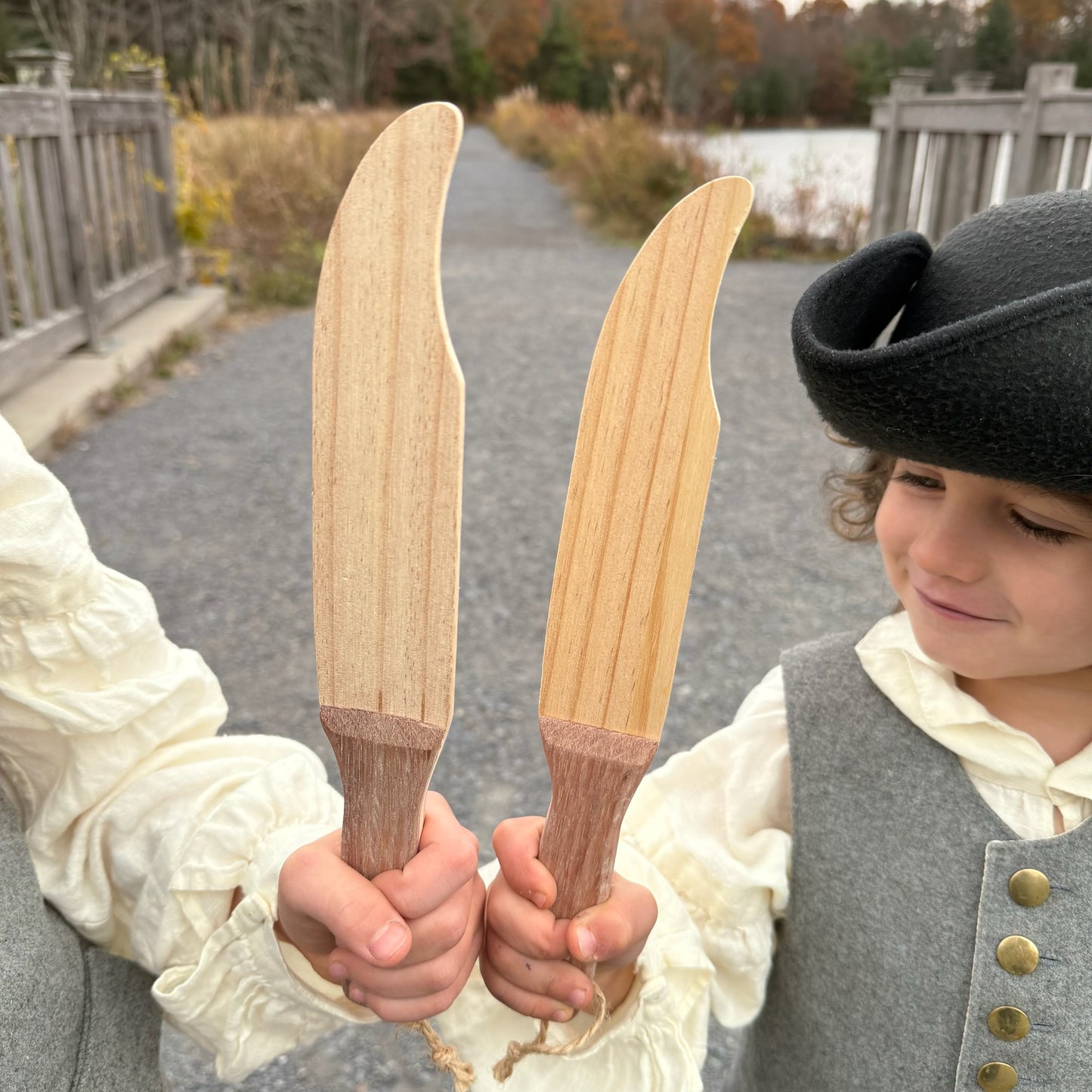 Two children in colonial attire holding wooden toy knives outdoors.