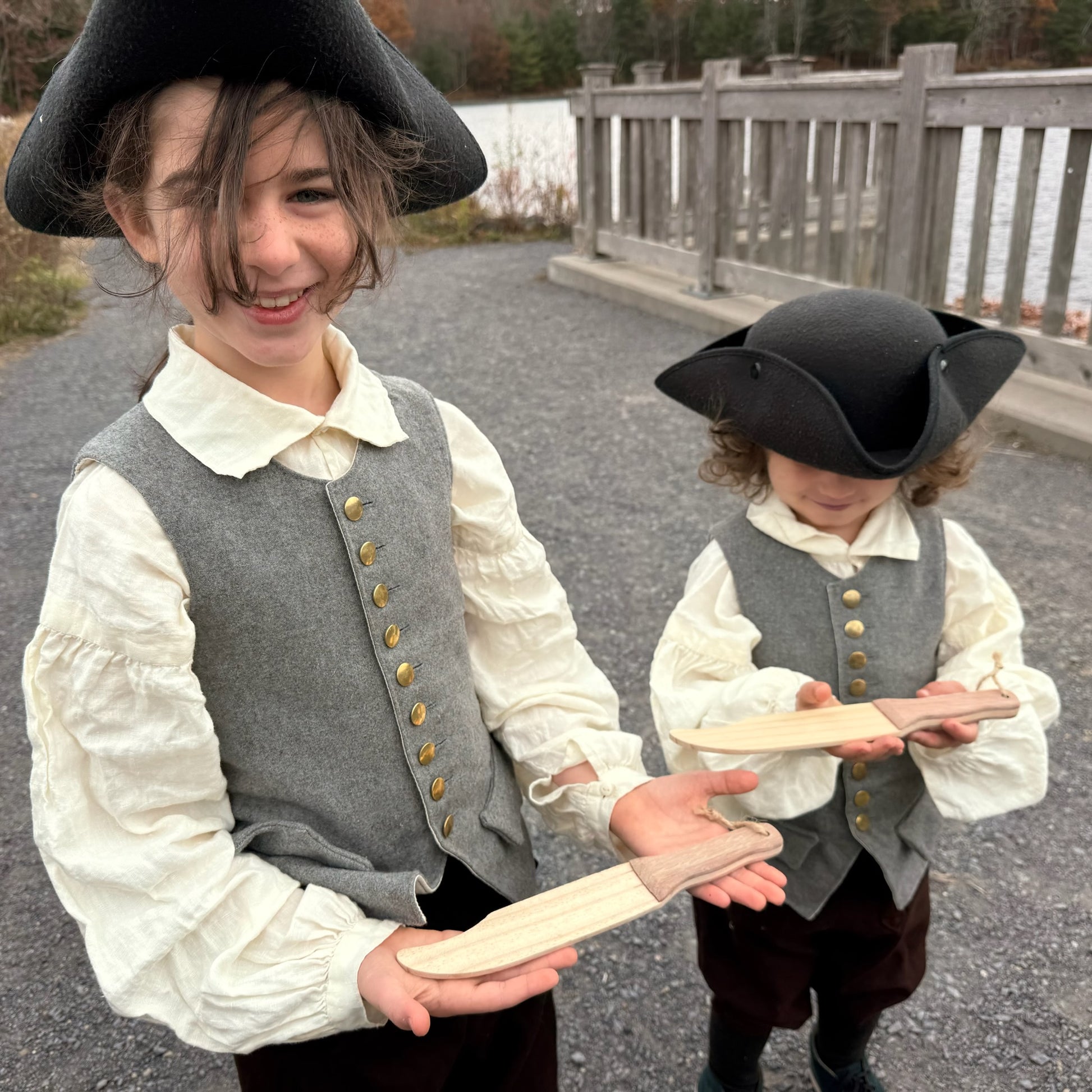 Two children in colonial-style clothing holding wooden toy knives outdoors.