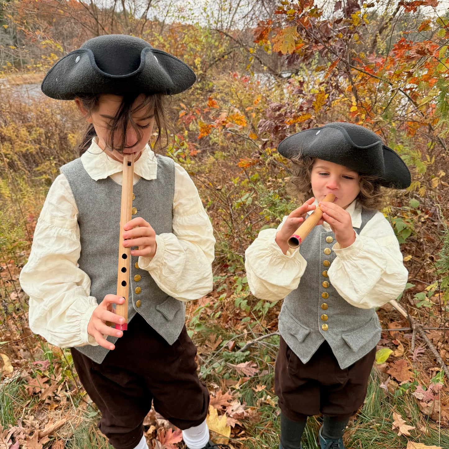 Two children in colonial-style clothing playing bamboo penny whistles outdoors in autumn.