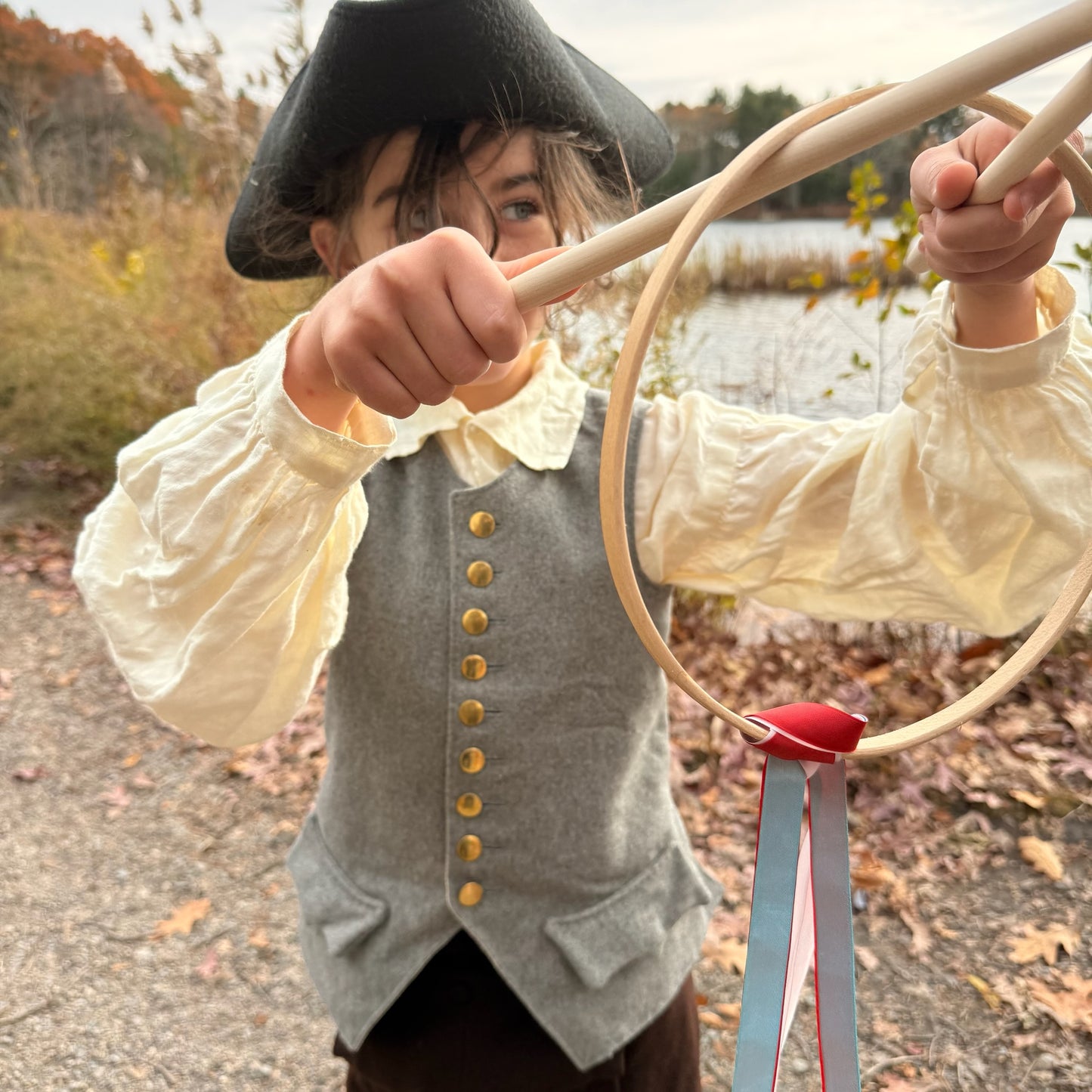 Child in colonial attire holding a Graces game outdoors near a lake.