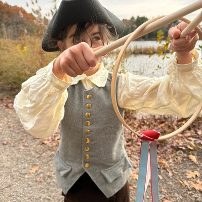 Child in colonial attire holding a Graces game outdoors near a lake.