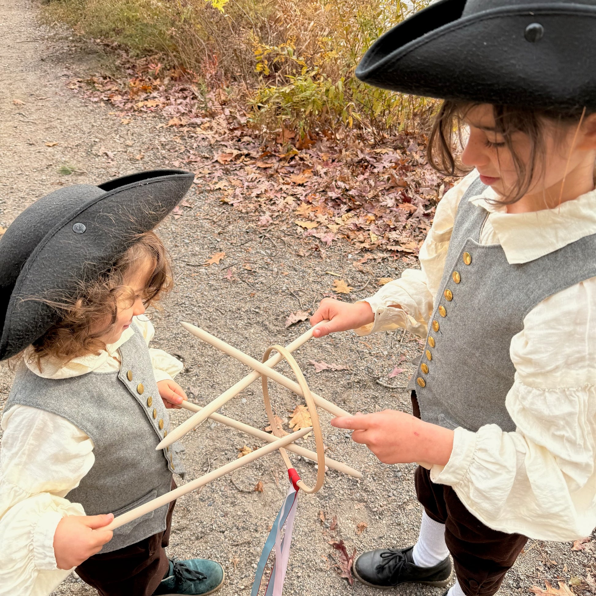 Two children in colonial attire playing with Graces game on a path with autumn leaves.