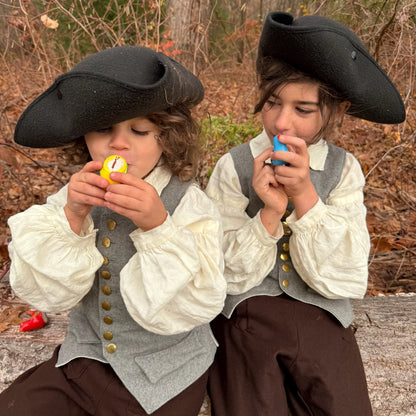 Two children in colonial attire blowing bird whistles outdoors.