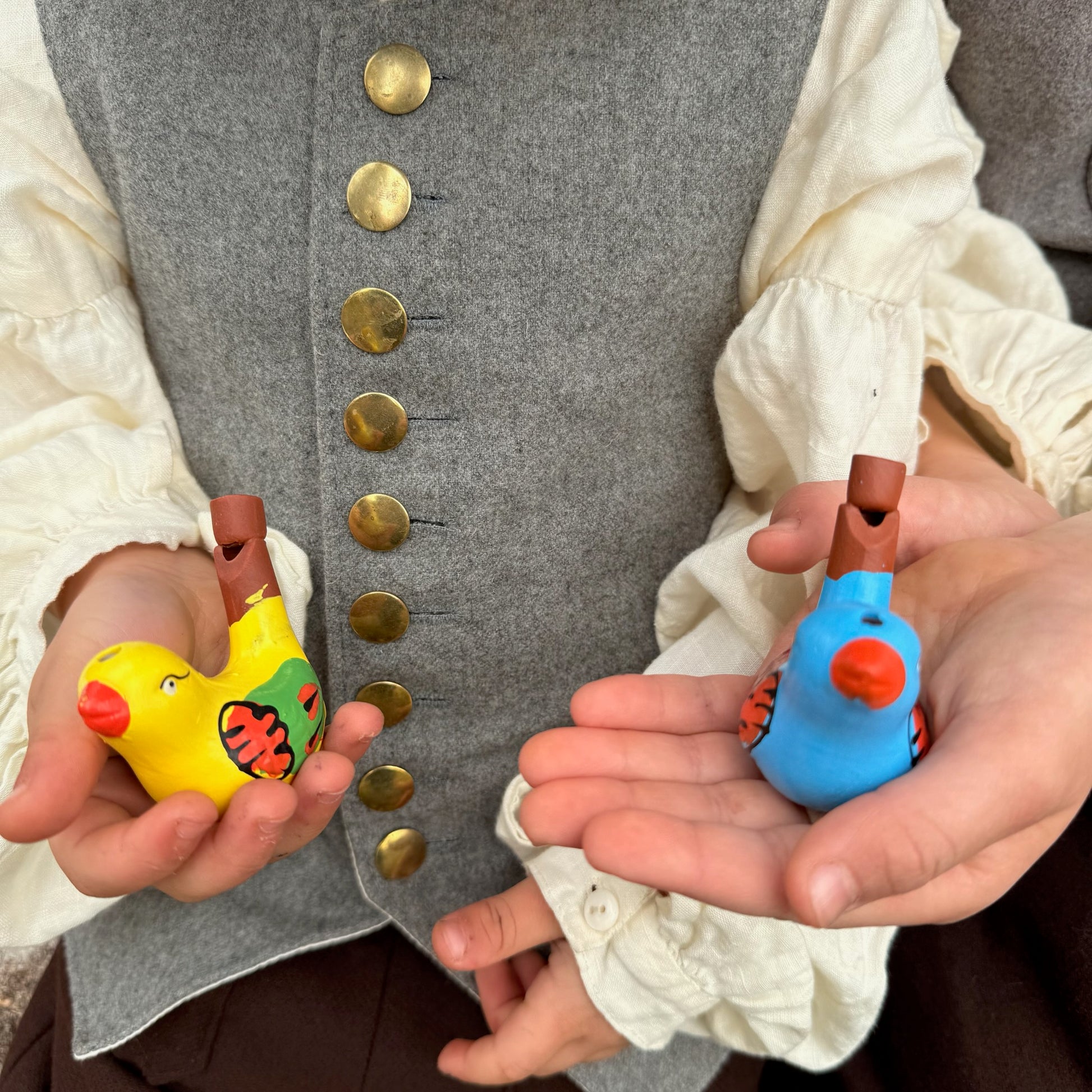 Two children wearing historical clothing holding colorful bird whistles.