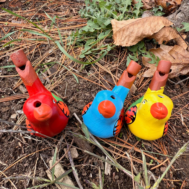 Three colorful bird-shaped whistles on the ground with leaves and twigs.