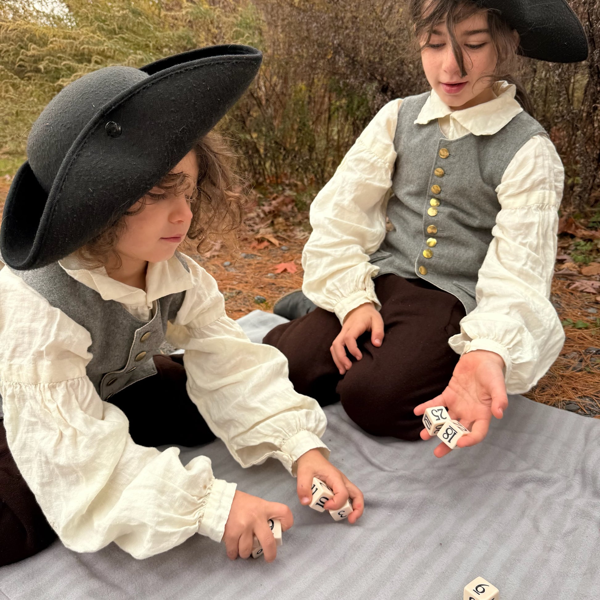 Two children in historical costumes playing with dice outdoors.