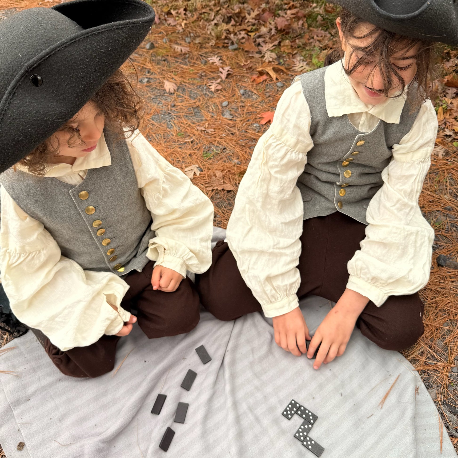 Two children in colonial attire sitting on a blanket outdoors playing with dominoes.