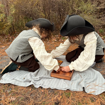 Two children in colonial attire playing with a wooden Ludo box outdoors.