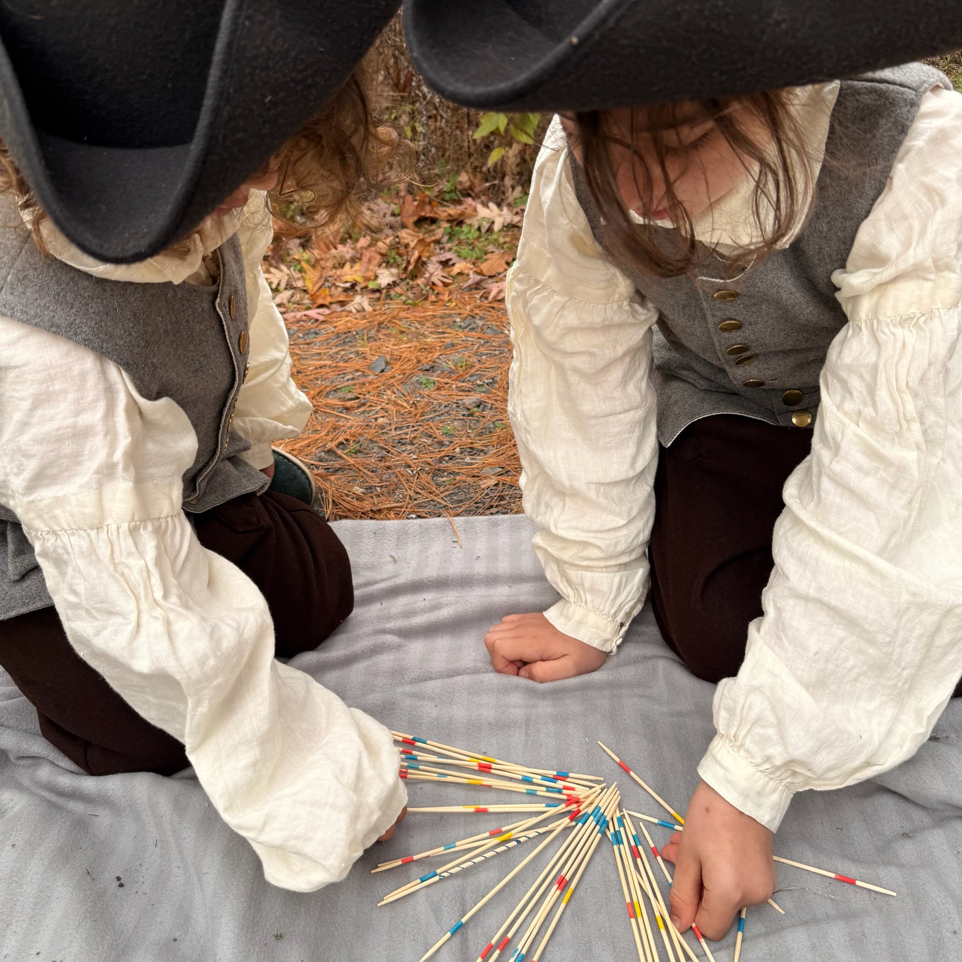 Two children in colonial attire playing with Pick Up Sticks on a blanket outdoors.