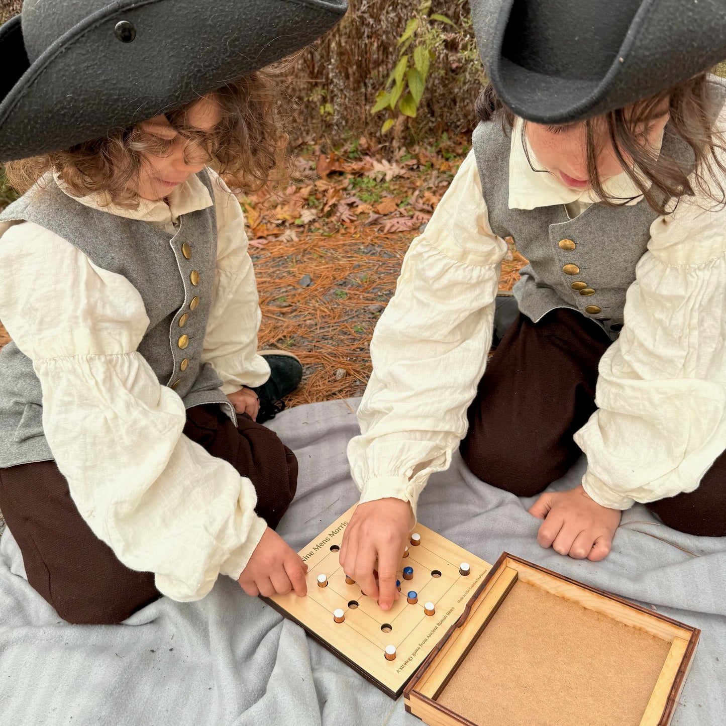 Two children in colonial attire playing a game of Nine Men's Morris outdoors.