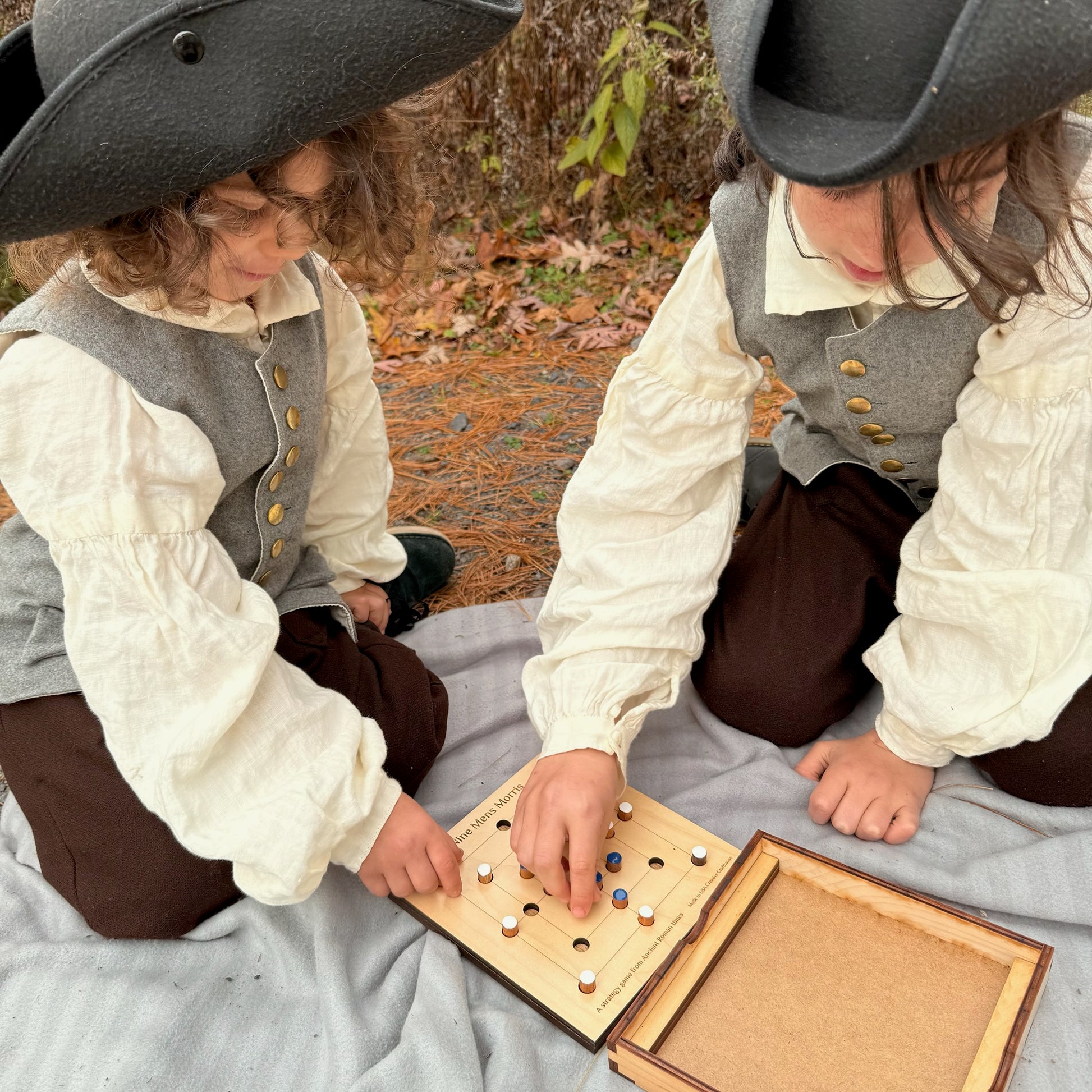 Two children in colonial attire playing a game of Nine Men's Morris outdoors.