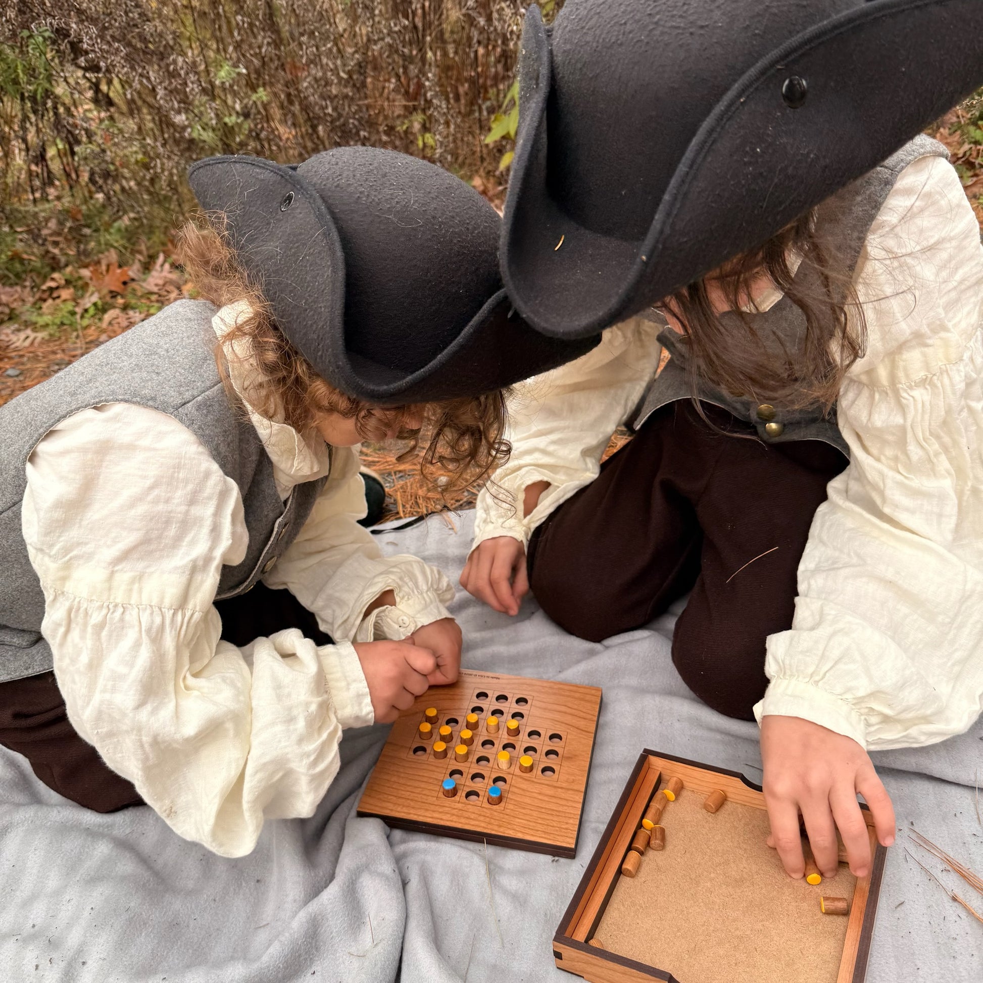 Two children in colonial attire playing a game of Solitaire outdoors.