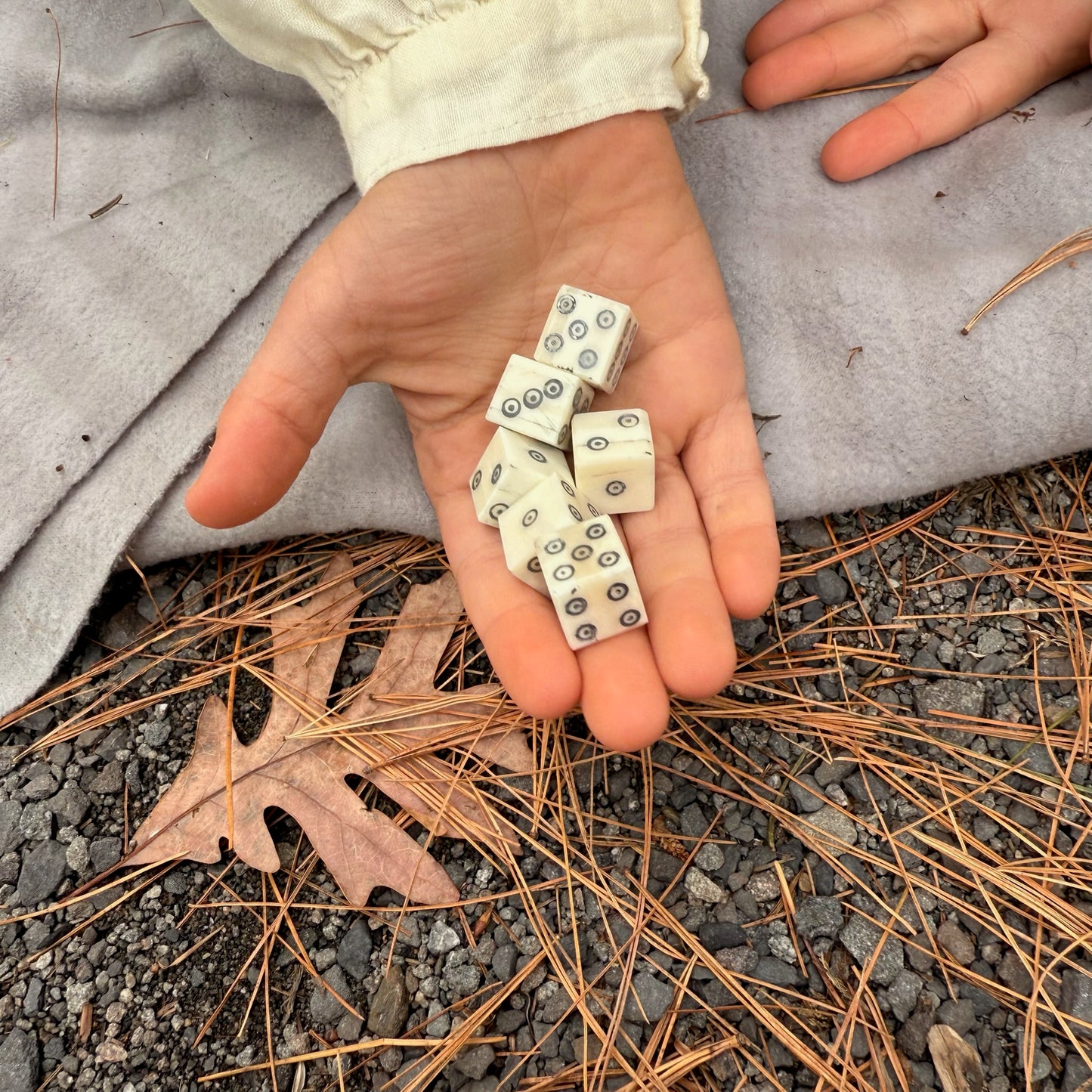 Hand holding small bone dice on a ground with leaves and fabric