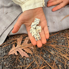 Hand holding small bone dice on a ground with leaves and fabric