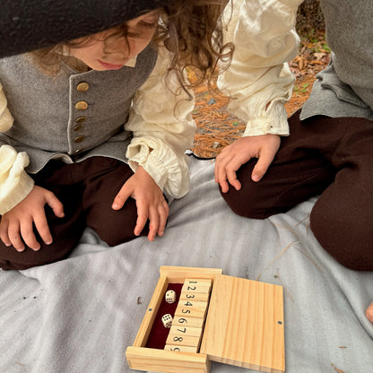 Two children in historical attire playing with a miniature Shut the Box game outdoors.