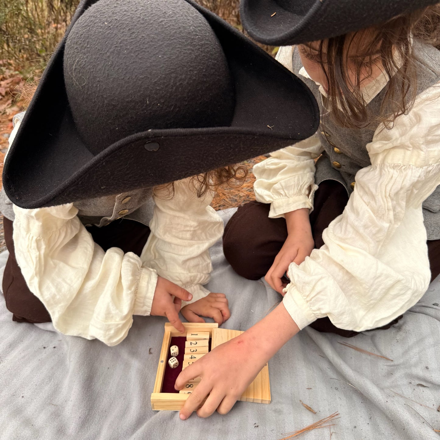 Two children in colonial attire playing with a miniature wooden Shut the Box game outdoors.