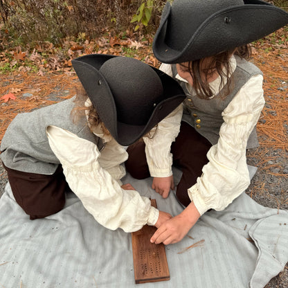 Two children in colonial attire sitting on the ground outdoors playing Cribbage.