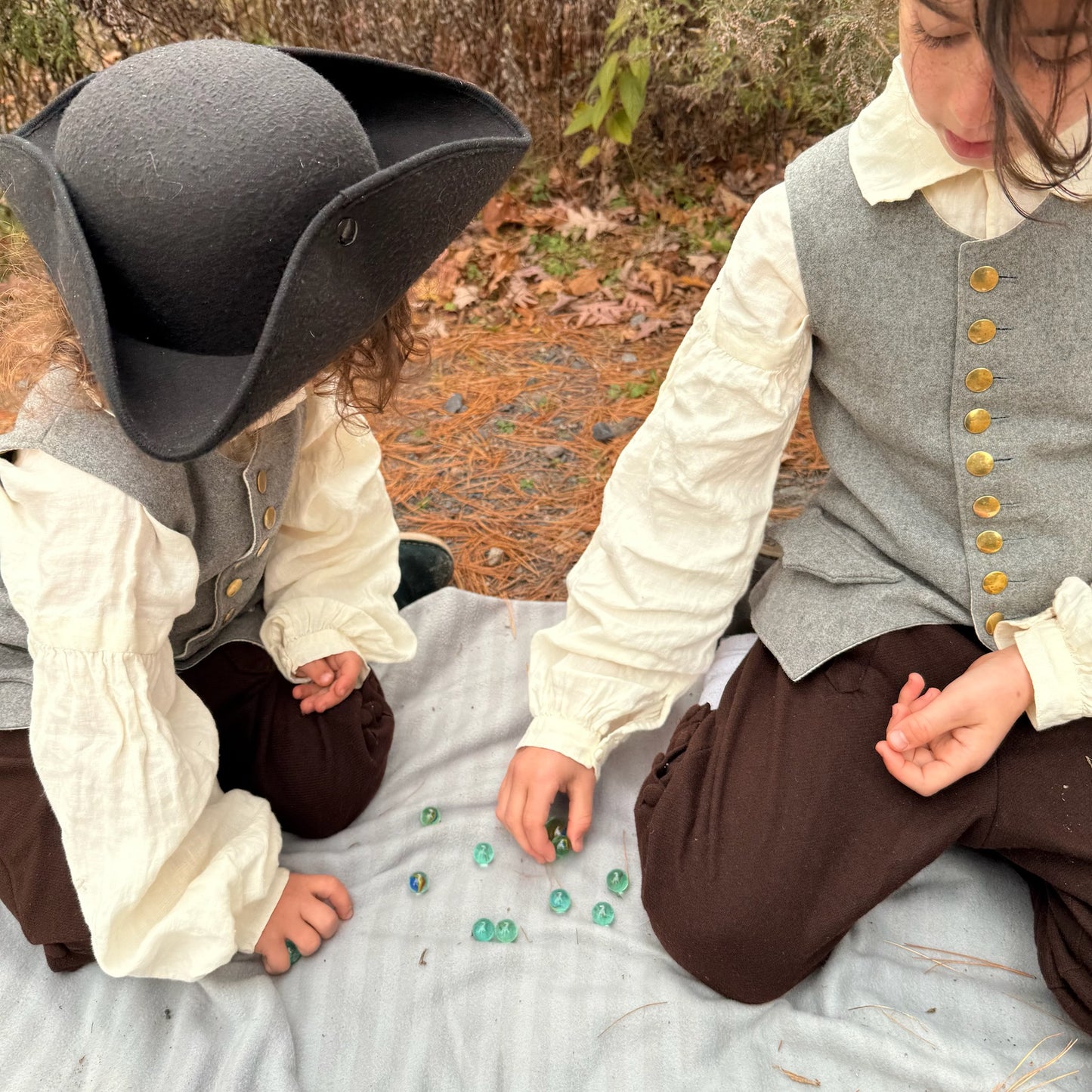 Two children in colonial attire playing with marbles outdoors.