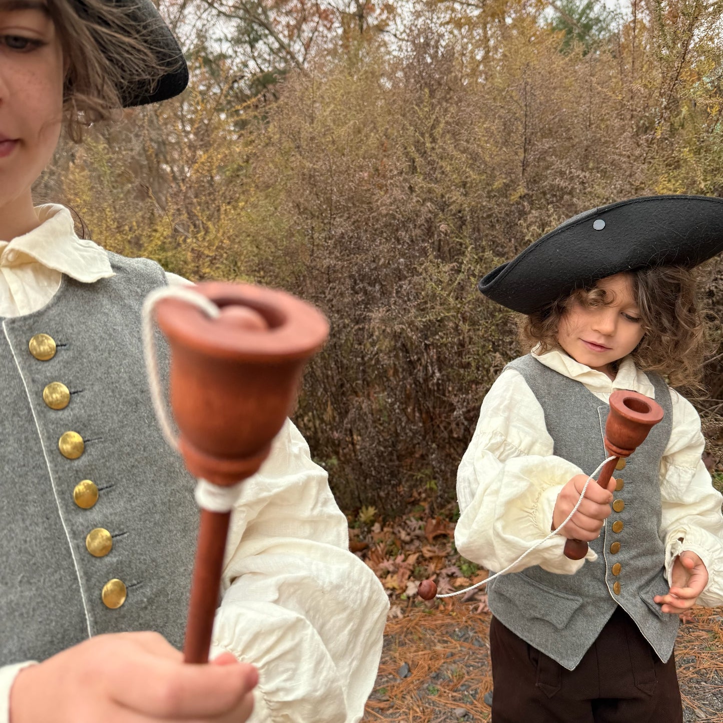 Two children in colonial attire playing with wooden cup and ball toys outdoors.