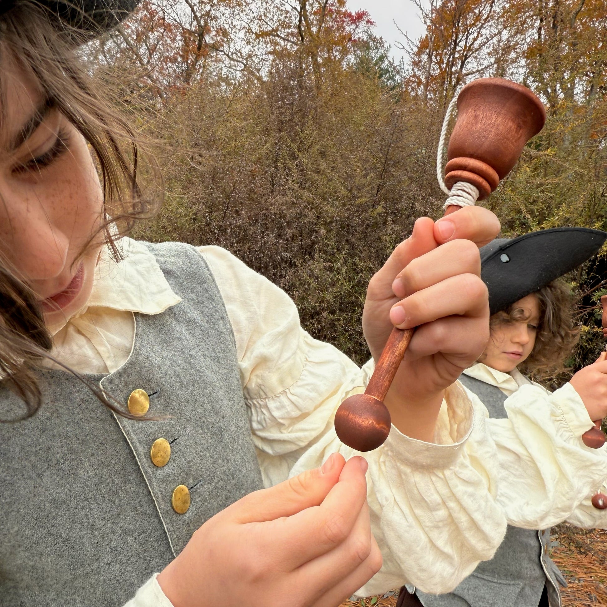 Two children in historical attire holding wooden cup and ball toys with trees in the background