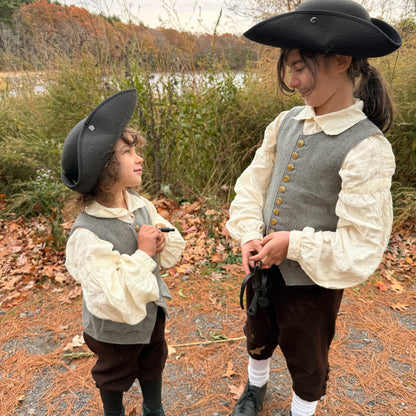 Two children in colonial-style clothing standing outdoors holding black iron shackles with a natural background.