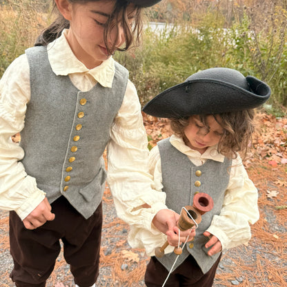 Two children in colonial-style clothing standing outdoors with a natural background and playing with cup and ball toys.