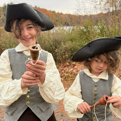 Two children in colonial attire holding wooden cup and ball toys outdoors with a forest background.