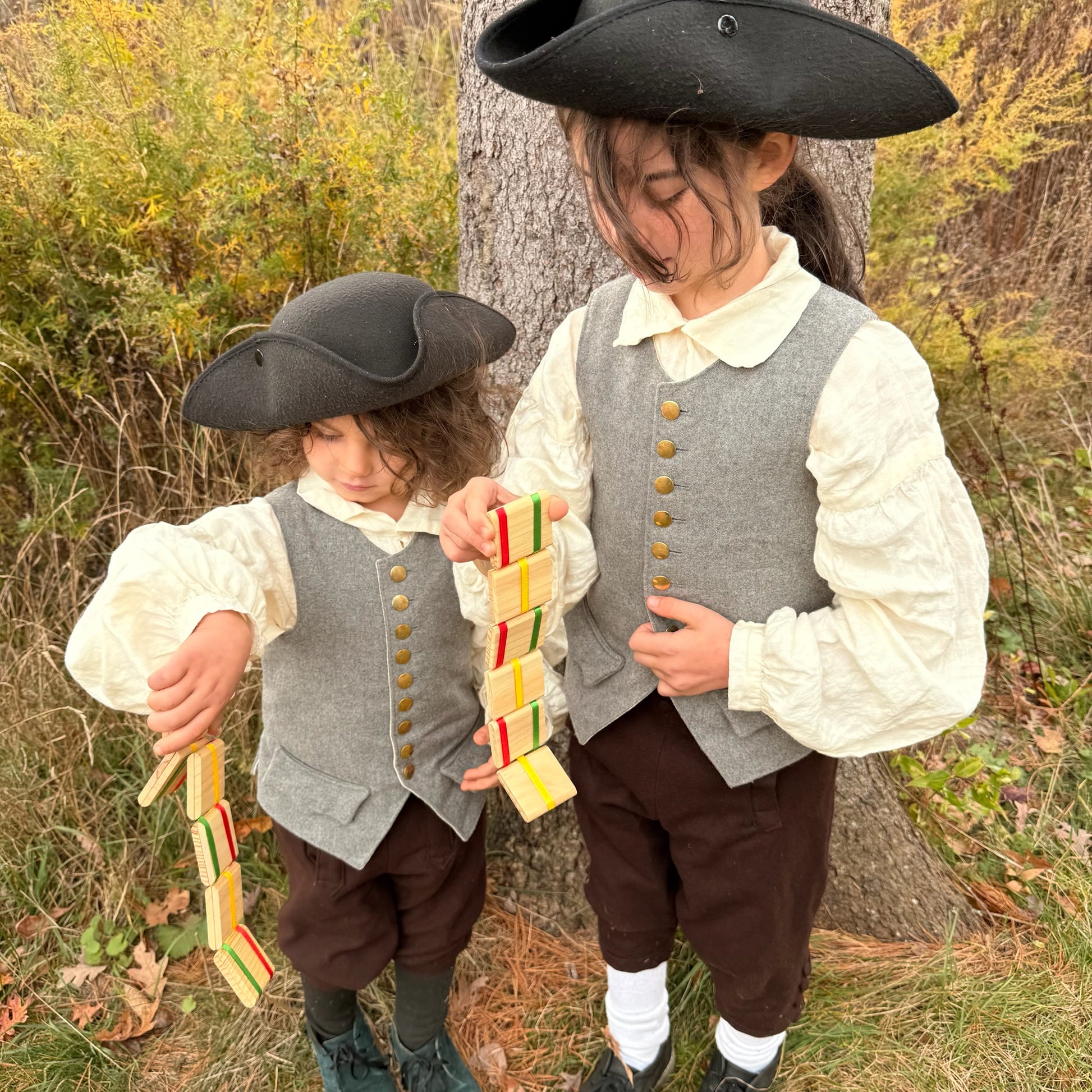Two children in colonial-style clothing standing outdoors near a tree playing with Jacob's Ladder toys.