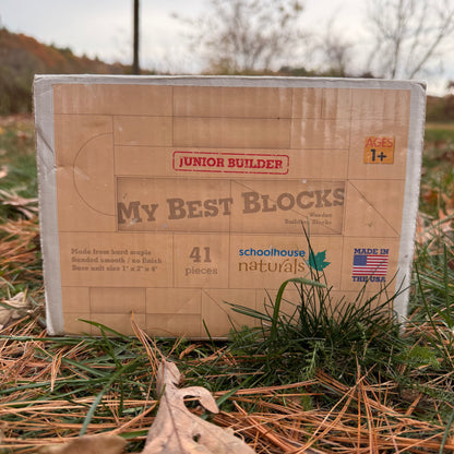 Cardboard box of 'Junior Builder My Best Blocks' wooden building blocks by Schoolhouse Naturals on a grass surface.