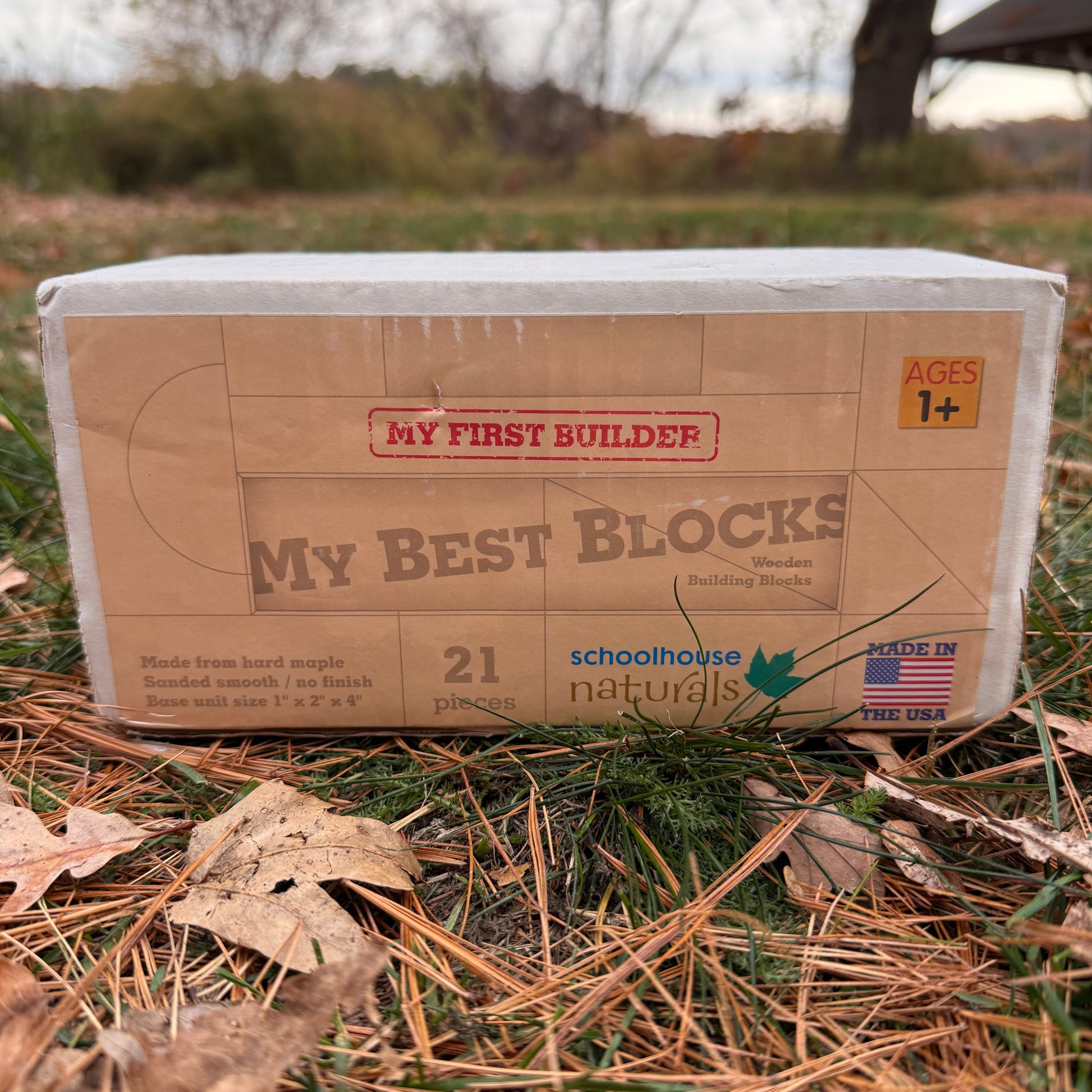 Cardboard box of 'My First Builder My Best Blocks' wooden building blocks by Schoolhouse Naturals on a grass surface.